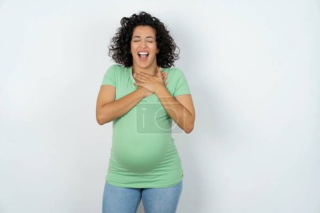 Foto de Mujer embarazada expresa felicidad, ríe agradablemente, mantiene las manos en el corazón - Imagen libre de derechos