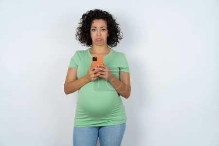 Foto de Mujer embarazada molesto utiliza aplicación de software móvil y navega información en Internet, sostiene teléfono móvil moderno - Imagen libre de derechos