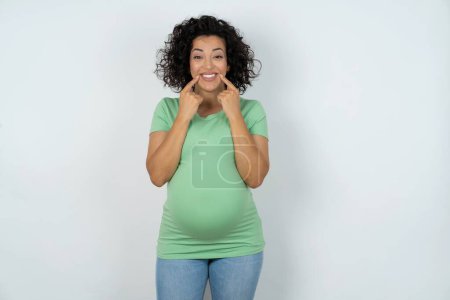 Foto de Mujer embarazada feliz con sonrisa dentada, mantiene los dedos índice cerca de la boca, los dedos señalando y forzando la sonrisa alegre - Imagen libre de derechos