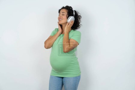 Photo for Pregnant woman with headphones on her head, listens to music, enjoying favourite song with closed eyes, holding hands on headset. - Royalty Free Image