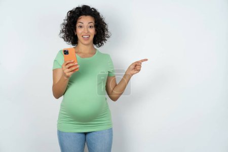 Téléchargez les photos : Femme enceinte étonnée tenant son téléphone et pointant du doigt à côté de l'espace de copie vide - en image libre de droit