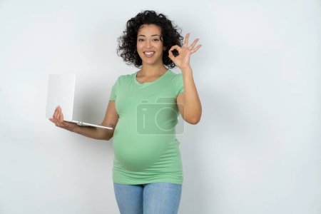 Téléchargez les photos : Femme enceinte positive tient netbook sans fil et montre okey symbole - en image libre de droit