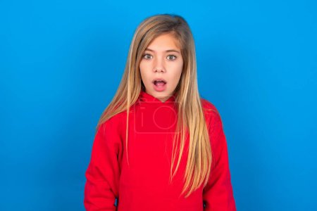 Foto de Hermosa chica adolescente caucásica con suéter rojo sobre la pared azul de haber aturdido y sorprendido mirada, con la boca abierta y la mandíbula cayó exclamando: Wow, no puedo creer esto. Sorpresa y shock - Imagen libre de derechos