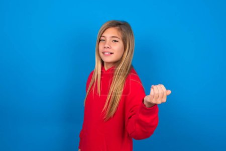 Foto de Hermosa chica adolescente caucásica con suéter rojo sobre la pared azul invitándole a venir, confiado y sonriente haciendo un gesto con la mano, siendo positivo y amigable. - Imagen libre de derechos