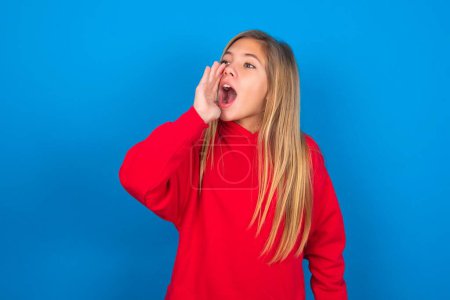 Photo for Beautiful caucasian teen girl wearing red sweater over blue wall profile view, looking happy and excited, shouting and calling to copy space. - Royalty Free Image