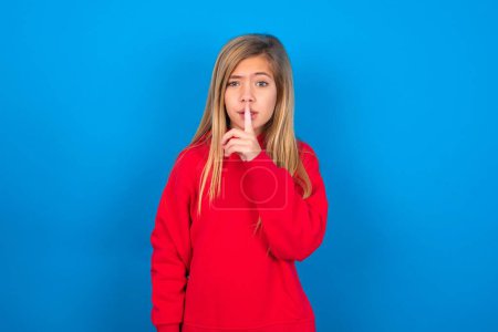 Téléchargez les photos : Belle adolescente caucasienne portant un pull rouge sur un mur bleu fait un geste de silence, demande silence. Ne dites pas mon secret ou ne parlez pas trop fort, s'il vous plaît.! - en image libre de droit