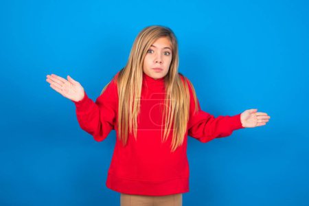 Photo for Puzzled and clueless beautiful caucasian teen girl wearing red sweater over blue wall with arms out, shrugging shoulders, saying: who cares, so what, I don't know. - Royalty Free Image