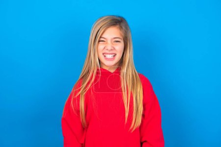 Foto de Loca loca hermosa chica adolescente caucásica usando suéter rojo sobre la pared azul aprieta los dientes airadamente, siendo molesto con el ruido que viene. Concepto de sentimiento negativo. - Imagen libre de derechos