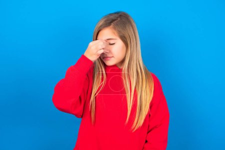 Foto de Muy molesto hermosa chica adolescente caucásica usando suéter rojo sobre la pared azul tocando la nariz entre los ojos cerrados, quiere llorar, tener una relación estresante o tener problemas con el trabajo - Imagen libre de derechos