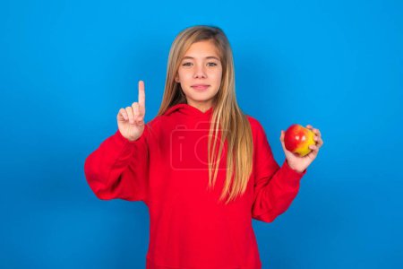 Foto de Hermosa chica adolescente caucásica usando suéter rojo sobre la pared azul mostrando y señalando hacia arriba con los dedos número uno mientras sonríe confiado y feliz. - Imagen libre de derechos