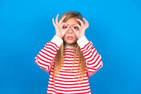 Photo for Playful excited beautiful caucasian teen girl wearing striped shirt over blue studio background showing Ok sign with both hands on eyes, pretending to wear spectacles. - Royalty Free Image