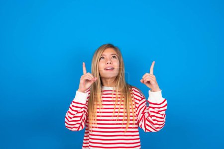 Photo for Successful friendly looking beautiful caucasian teen girl wearing striped shirt over blue studio background exclaiming excitedly, pointing both index fingers up, indicating something. - Royalty Free Image
