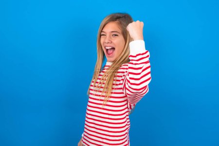 Photo for Overjoyed beautiful caucasian teen girl wearing striped shirt over blue studio background glad to receive good news, clenching fist and making winning gesture. - Royalty Free Image