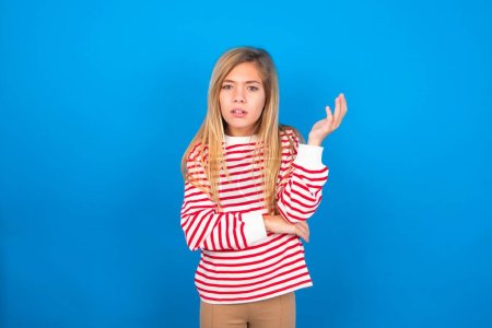 Foto de Studio shot of frustrated beautiful caucasian teen girl wearing striped shirt over blue studio background gesturing with raised palm, frowning, being displeased and confused with dumb question. - Imagen libre de derechos