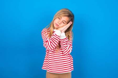 Photo for Relax and sleep time. Tired beautiful caucasian teen girl wearing striped shirt over blue studio background with closed eyes leaning on palms making sleeping gesture. - Royalty Free Image