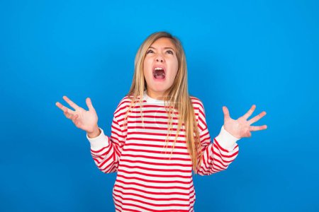 Photo for Beautiful caucasian teen girl wearing striped shirt over blue studio background crying and screaming. Human emotions, facial expression concept. Screaming, hate, rage. - Royalty Free Image