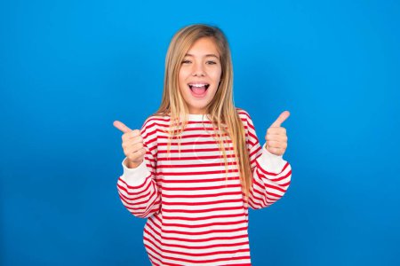 Téléchargez les photos : Optimiste belle adolescente caucasienne portant une chemise rayée sur fond de studio bleu montrant pouces vers le haut avec des émotions positives. Concept de qualité et de recommandation. - en image libre de droit