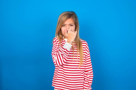 Téléchargez les photos : Belle caucasienne adolescent fille portant chemise rayée sur fond bleu studio tenant son nez en raison d'une mauvaise odeur. - en image libre de droit