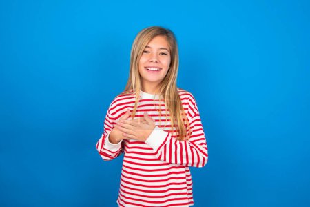 Photo for Honest beautiful caucasian teen girl wearing striped shirt over blue studio background keeps hands on chest, touched by compliment or makes promise, looks at camera with great pleasure. - Royalty Free Image
