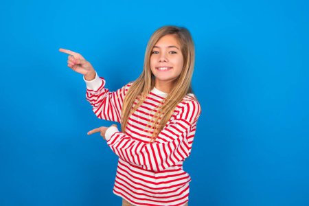 beautiful caucasian teen girl wearing striped shirt over blue studio background points aside with  surprised expression with mouth opened, shows something amazing. Advertisement concept.