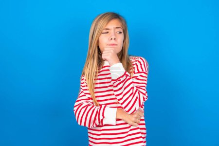 Photo for Beautiful caucasian teen girl wearing striped shirt over blue studio background with hand under chin and looking sideways with doubtful and skeptical expression, suspect and doubt. - Royalty Free Image