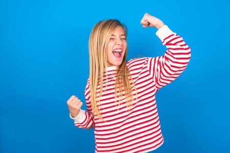 Téléchargez les photos : Attrayant belle caucasienne adolescent fille portant rayé chemise sur fond studio bleu célébrer une victoire frapper l'air avec ses poings et un sourire de dents rayonnantes - en image libre de droit