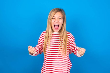Portrait of beautiful caucasian teen girl wearing striped shirt over blue studio background looks with excitement at camera, keeps hands raised over head, notices something unexpected reacts on sudden news.