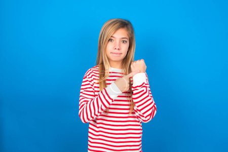 Téléchargez les photos : Belle fille adolescente caucasienne portant une chemise rayée sur fond bleu studio à la hâte pointant vers regarder le temps, l'impatience, contrarié et en colère pour retard de la date limite - en image libre de droit