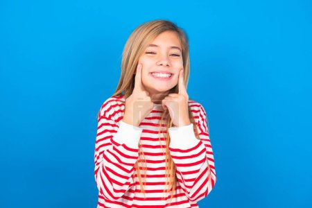 Photo for Strong healthy straight white teeth. Close up portrait of happy beautiful caucasian teen girl wearing striped shirt over blue studio background with beaming smile pointing on perfect clear white teeth. - Royalty Free Image