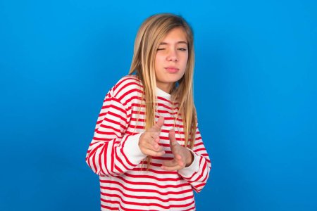 Photo for Hey you, bang. Joyful and charismatic good-looking beautiful caucasian teen girl wearing striped shirt over blue studio background winking and pointing with finger pistols at camera happily and cheeky. - Royalty Free Image