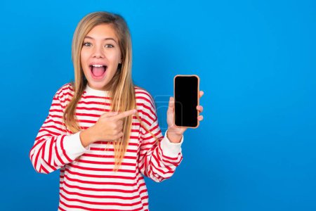 Photo for Excited beautiful caucasian teen girl wearing striped shirt over blue studio background holding and pointing with finger at smartphone with blank screen. Advertisement concept. - Royalty Free Image