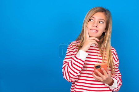 Foto de Imagen de una niña adolescente caucásica soñando con un pensamiento que lleva una camisa a rayas sobre un fondo azul del estudio usando un teléfono móvil y sosteniendo la mano en la cara. Toma de decisiones y concepto de redes sociales. - Imagen libre de derechos