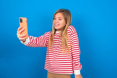 Foto de Foto aislada de alegre alegre hermosa chica adolescente caucásica con camisa a rayas sobre fondo de estudio azul, hace selfie con teléfono móvil. Concepto de personas, tecnología y ocio - Imagen libre de derechos