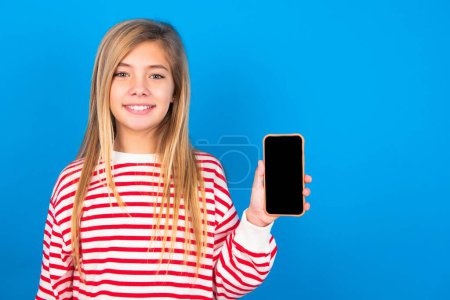 Foto de Sonriente hermosa chica adolescente caucásica con camisa a rayas sobre fondo de estudio azul que muestra la pantalla del teléfono vacío. Concepto de publicidad y comunicación. - Imagen libre de derechos