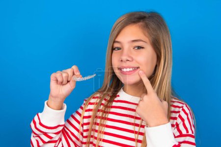 belle adolescente caucasienne portant une chemise rayée sur fond de studio bleu tenant un aligneur invisible et pointant vers ses dents droites parfaites. Concept de soins dentaires et de confiance.