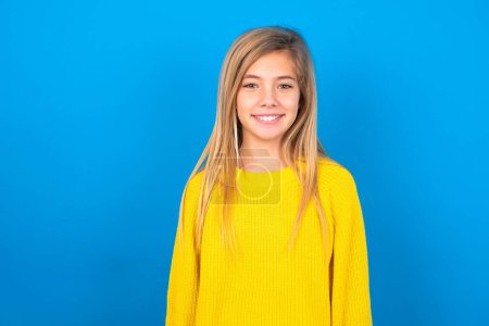 Téléchargez les photos : Belle jeune fille blonde portant un pull jaune sur le mur bleu avec un joli sourire rayonnant expression heureuse. Concept des émotions positives - en image libre de droit