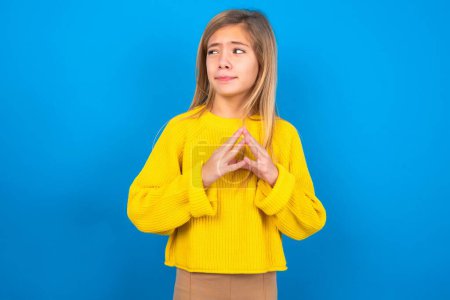 Photo for Beautiful blonde teen girl wearing yellow sweater over blue wall steepled fingers and looks mysterious aside has great evil plan in mind - Royalty Free Image