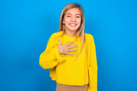 Photo for Beautiful blonde teen girl wearing yellow sweater over blue wall smiles toothily cannot believe eyes expresses good emotions and surprisement - Royalty Free Image