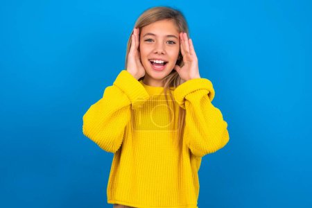Photo for Beautiful blonde teen girl wearing yellow sweater over blue wall Pleasant looking cheerful, Happy reaction - Royalty Free Image