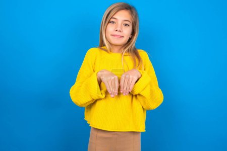 Photo for Beautiful blonde teen girl wearing yellow sweater over blue wall makes bunny paws and looks with innocent expression plays with her little kid - Royalty Free Image