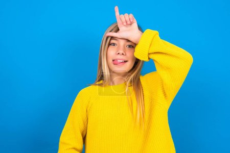 Foto de Hermosa rubia adolescente chica usando amarillo suéter sobre azul gestos de la pared con el dedo en la frente hace gesto perdedor se burla de la gente muestra la lengua - Imagen libre de derechos