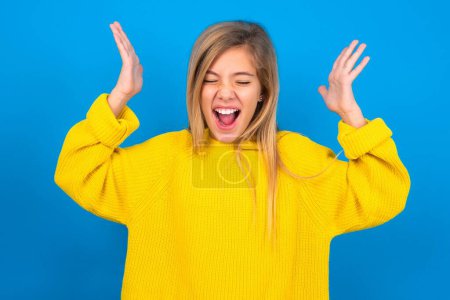 Photo for Beautiful blonde teen girl wearing yellow sweater over blue wall goes crazy as head goes around feels stressed because of horrible situation - Royalty Free Image