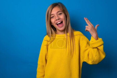 Photo for Beautiful blonde teen girl wearing yellow sweater over blue wall foolishness around shoots in temple with fingers makes suicide gesture. Funny model makes finger gun pistol - Royalty Free Image