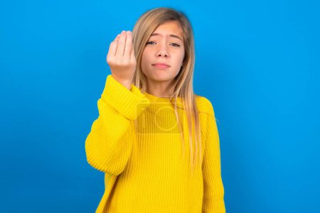 Foto de Hermosa rubia adolescente chica vistiendo suéter amarillo sobre la pared azul haciendo gesto italiano con la mano y los dedos expresión de confianza - Imagen libre de derechos