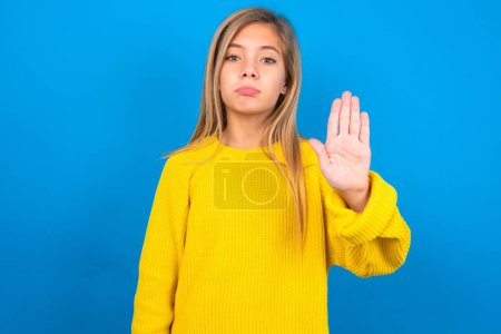 Photo for Beautiful blonde teen girl wearing yellow sweater over blue wall shows stop sign prohibition symbol keeps palm forward to camera with strict expression - Royalty Free Image