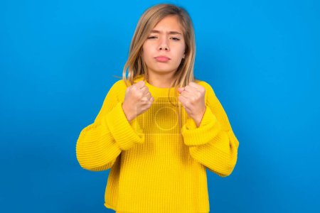 Foto de Displeased annoyed beautiful blonde teen girl wearing yellow sweater over blue wall clenches fists, gestures pissed, ready to revenge, looks with aggression at camera stands full of hate, being pressured - Imagen libre de derechos