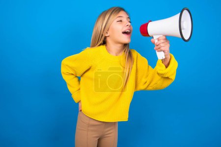 Foto de Divertido hermosa rubia adolescente con suéter amarillo sobre la pared azul Gente emociones sinceras concepto de estilo de vida. Prepara espacio para copias. Gritando en megáfono. - Imagen libre de derechos