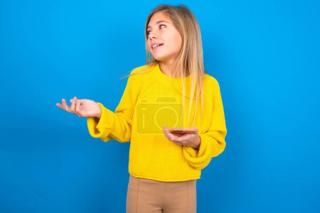 Photo for Happy pleased beautiful blonde teen girl wearing yellow sweater over blue wall raises palm and holds cellphone uses high speed internet for text messaging or video calls - Royalty Free Image