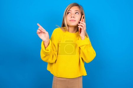 Photo for Beautiful blonde teen girl wearing yellow sweater over blue wall speaks on mobile phone spends free time indoors calls to friend. - Royalty Free Image