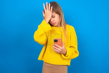 Photo for Upset depressed beautiful blonde teen girl wearing yellow sweater over blue wall makes face palm as forgot about something important holds mobile phone expresses sorrow and regret blames - Royalty Free Image